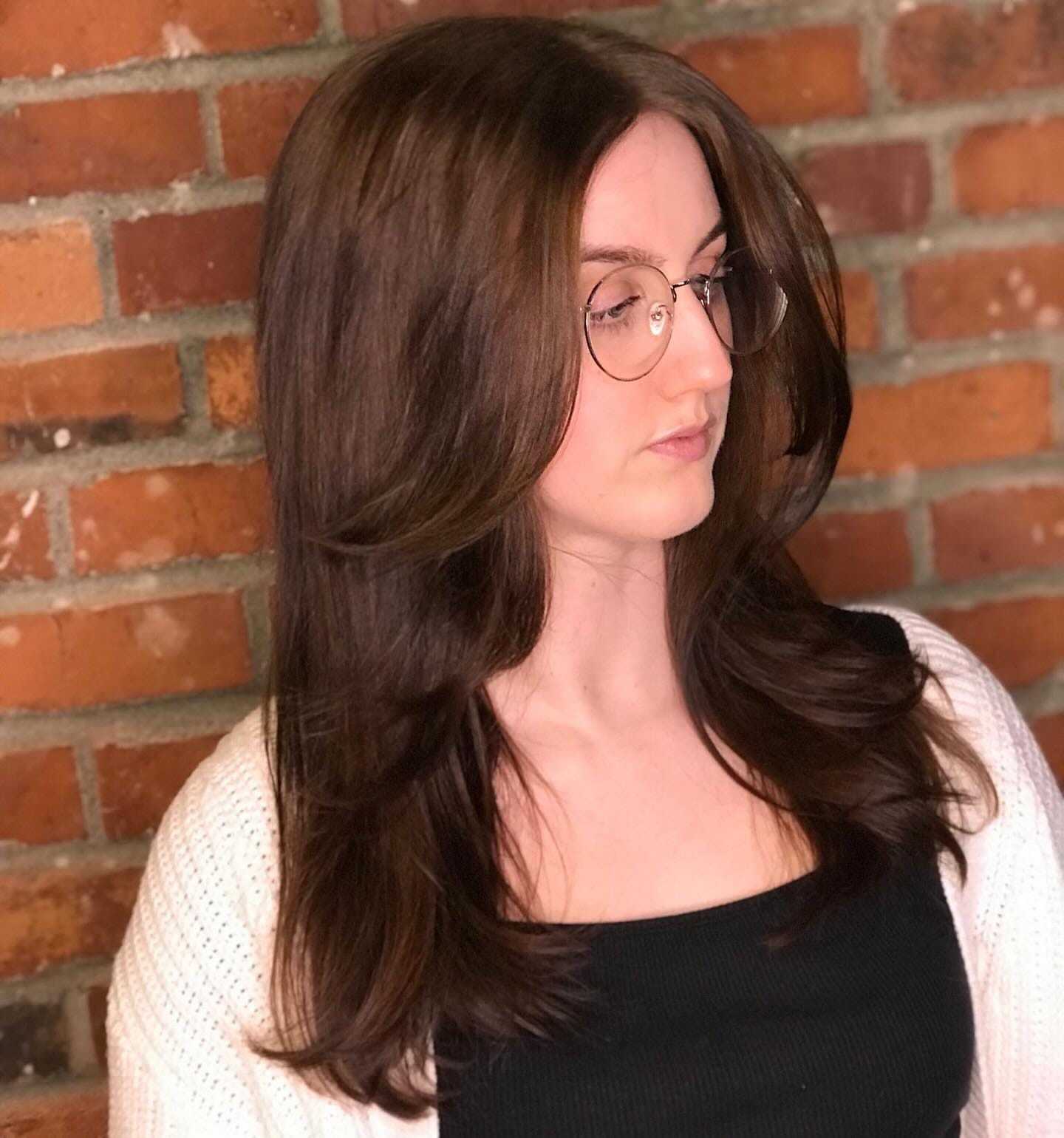 Woman with long brown hair and glasses, standing against a brick wall.