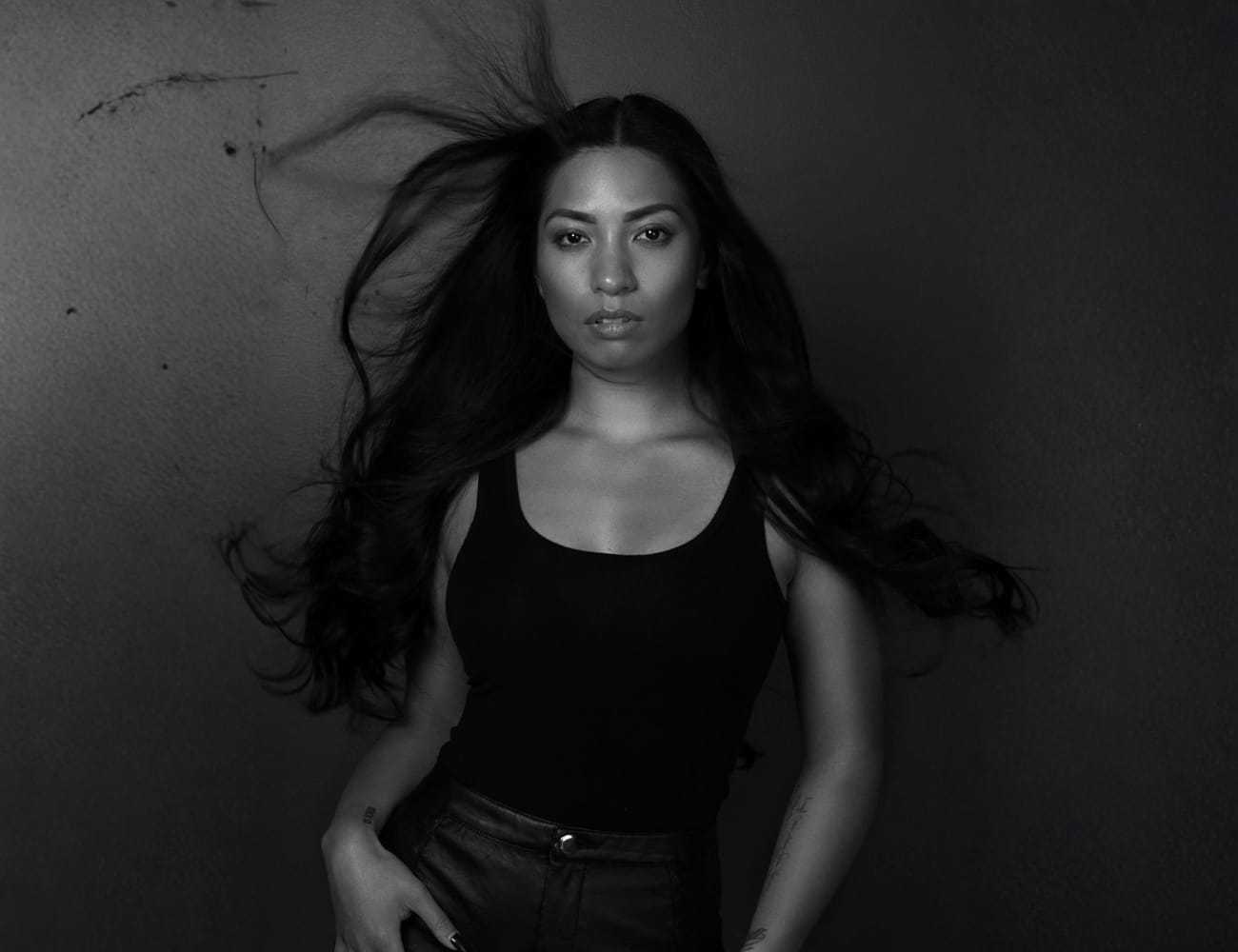 A woman with long hair blowing behind her, wearing a black top in a dramatic black-and-white photo.