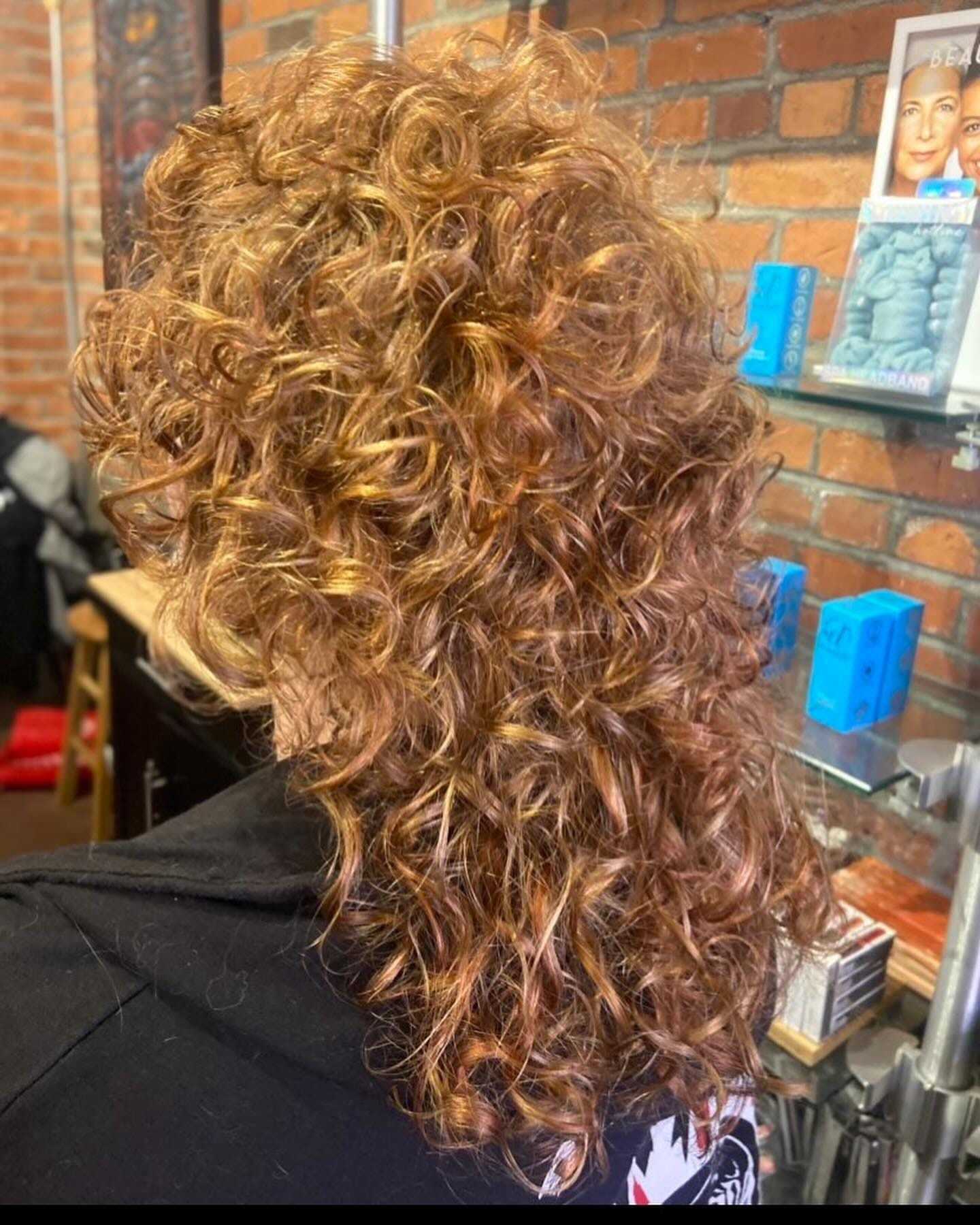 Curly auburn hair styled in a mullet, set against a brick wall background.